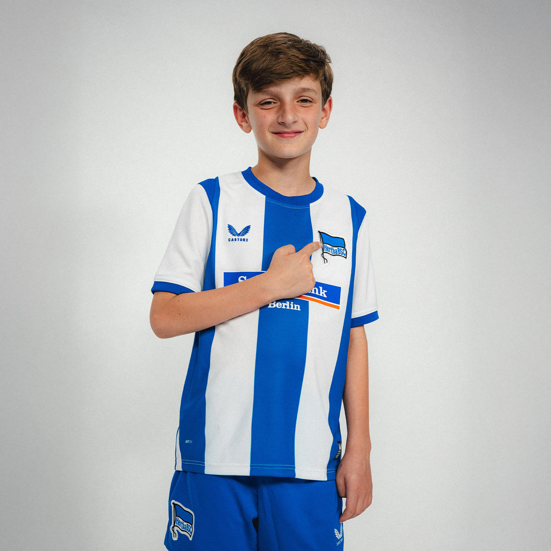 Hertha Heimtrikot Kids 26/27 Model zeigt auf Hertha Fahne